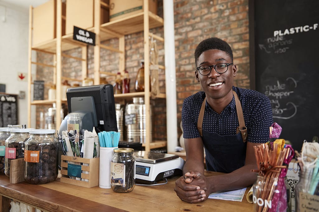 portrait of a black male in a black top with white polka dots standing behind the counter of a business or coffee shop