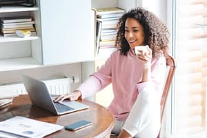 Black woman with curly hair and a pink sweater holding a white tea cup while freelancing at home on her laptop