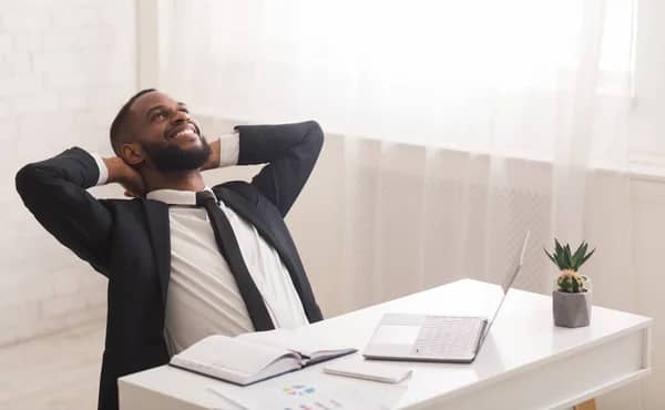 Relaxed black businessman in a black suit and black tie smiling and looking up in an office with a laptop notebook and phone on a white desktop