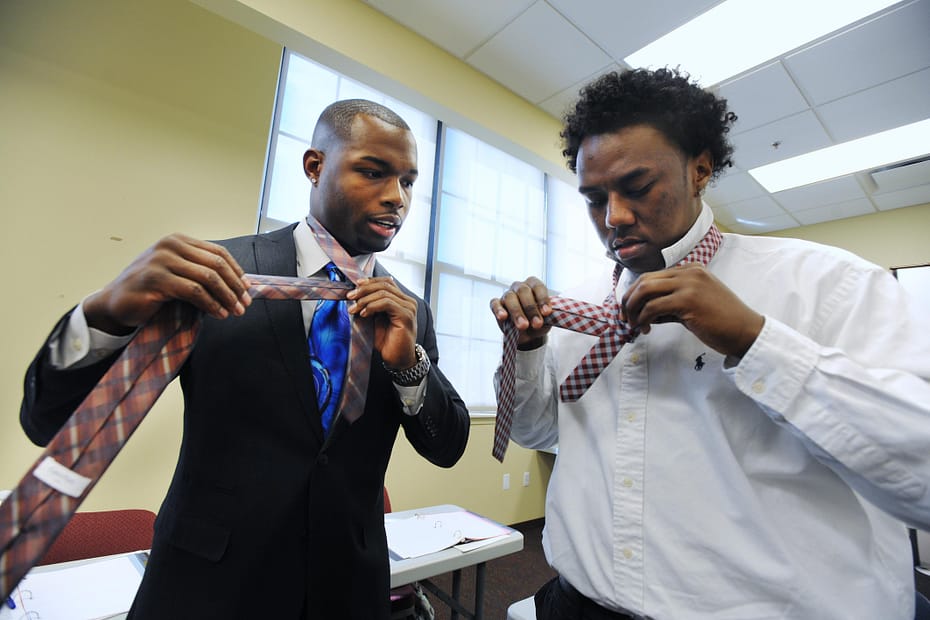 Black male in a black suit and blue tie showing another black male in a white shirt how to tie a tie as a mentor and mentee
