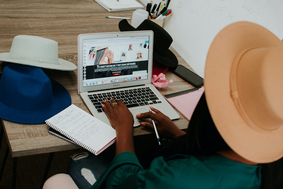 portrait of a Black woman with a green top and peach hat typing on a laptop with a grey pen and notebook and a variety of other hats on a table as she looks at her online hat website