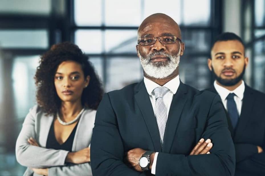 portrait of a black woman in a silver blazer and black shirt holding her arms with two black men in black suits with their arms folded looking powerful and successful in an office or business setting