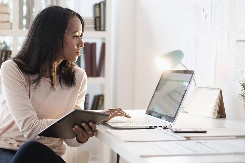 portrait of a Black woman in a light top and dark bottoms sitting at a desk in an office with a tablet in one hand her other hand typing on a laptop