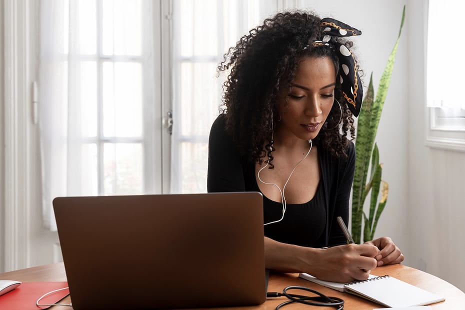 Black woman sitting down with a pen and notebook while listening to music and working at home on her laptop