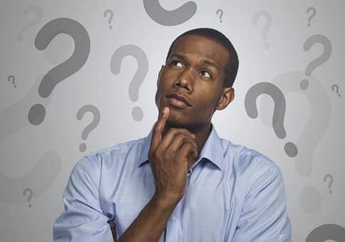 portrait of a black man thinking with a light blue shirt and arms folding with a finger on his chin. Question marks make up the background of the image.