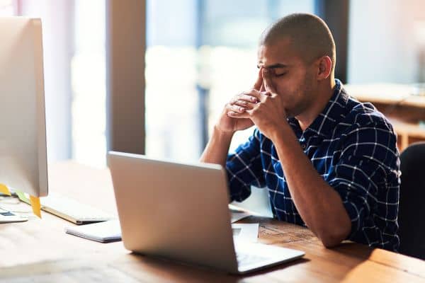 portrait of a frustrated or stressed black man sitting at desk with a laptop and his fingers touching his eyebrows