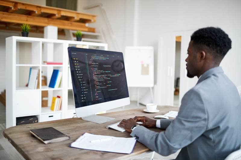 portrait of a black male in a blue top sitting at the desk typing or designing a website with code on his macbook in an office