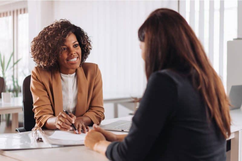 portrait of a black business woman in a brown blazer smiling in an office with a potential client or customer in front of her with red hair and a black top