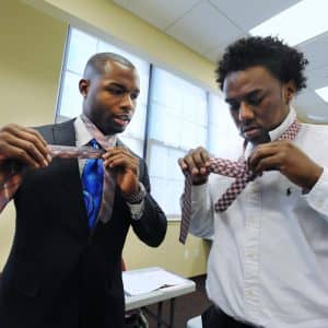 Black male in a black suit and blue tie showing another black male in a white shirt how to tie a tie as a mentor and mentee
